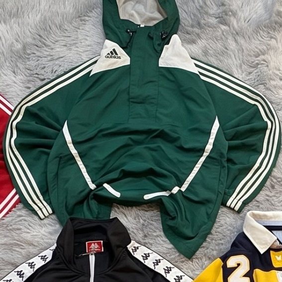 Anorak de tres rayas Adidas verde con capucha extendida sobre una alfombra gris